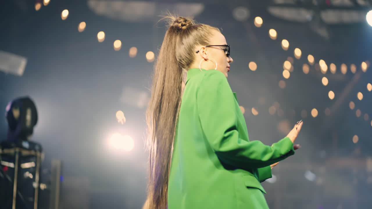una joven con una chaqueta verde baila en el escenario de un club nocturno.