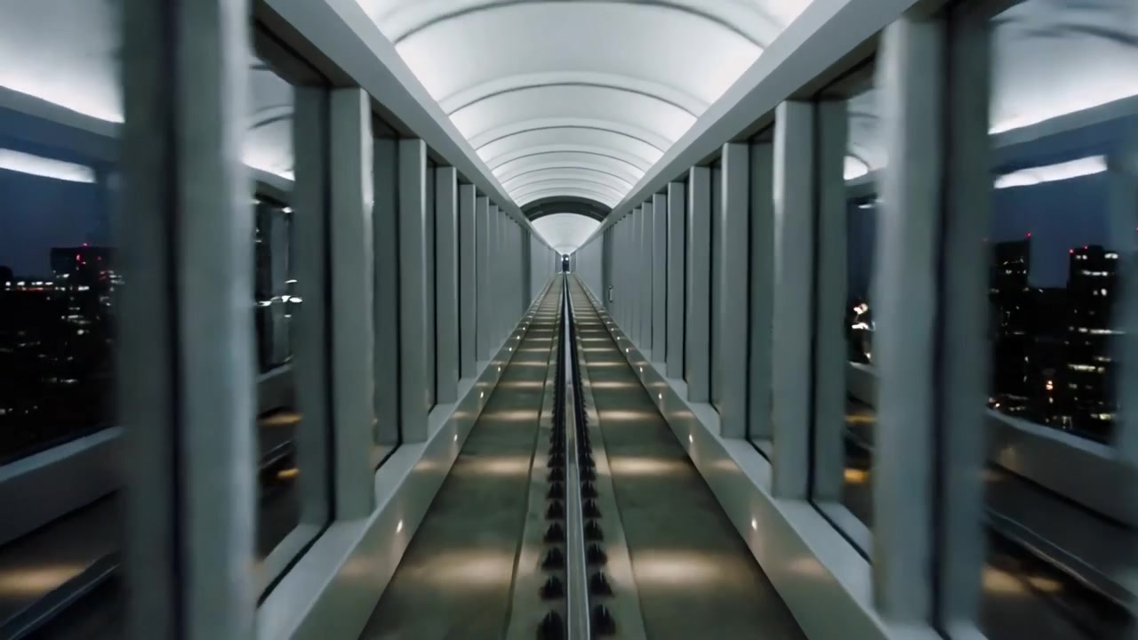 Telecamera che si muove attraverso un corridoio di vetro, rivelando una figura distante più avanti, con un binario centrale e luci.
