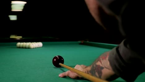 a brutal man with tattoos on his hands is playing russian billiards in a bar.