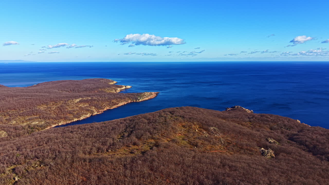 La costa incontra il mare blu sotto un cielo terso a mezzogiorno, con terra marrone e colline distanti visibili sullo sfondo