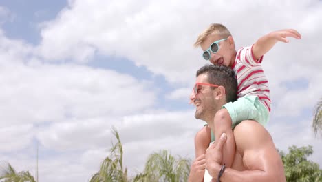 Portrait-of-happy-caucasian-father-carrying-his-son-with-sunglasses-at-beach-house