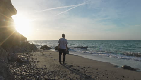 man walks on beach during sunny morning, stops, throws a stone, long shadow casts behind, sea waves wash onshore