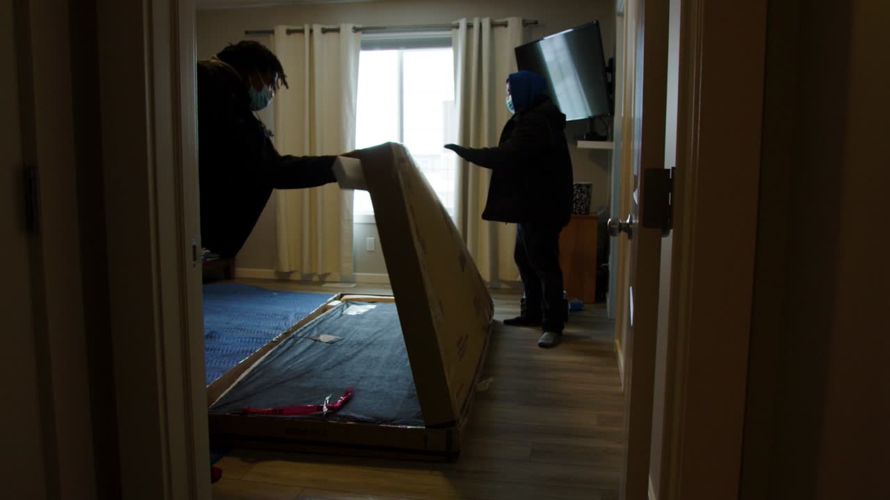 Shot through door of two delivery workers cutting tape and opening large box in bedroom light shines through window