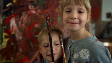 hijo alegre poniendo pincel en la nariz en el estudio de arte, hermana feliz mirando a la cámara.