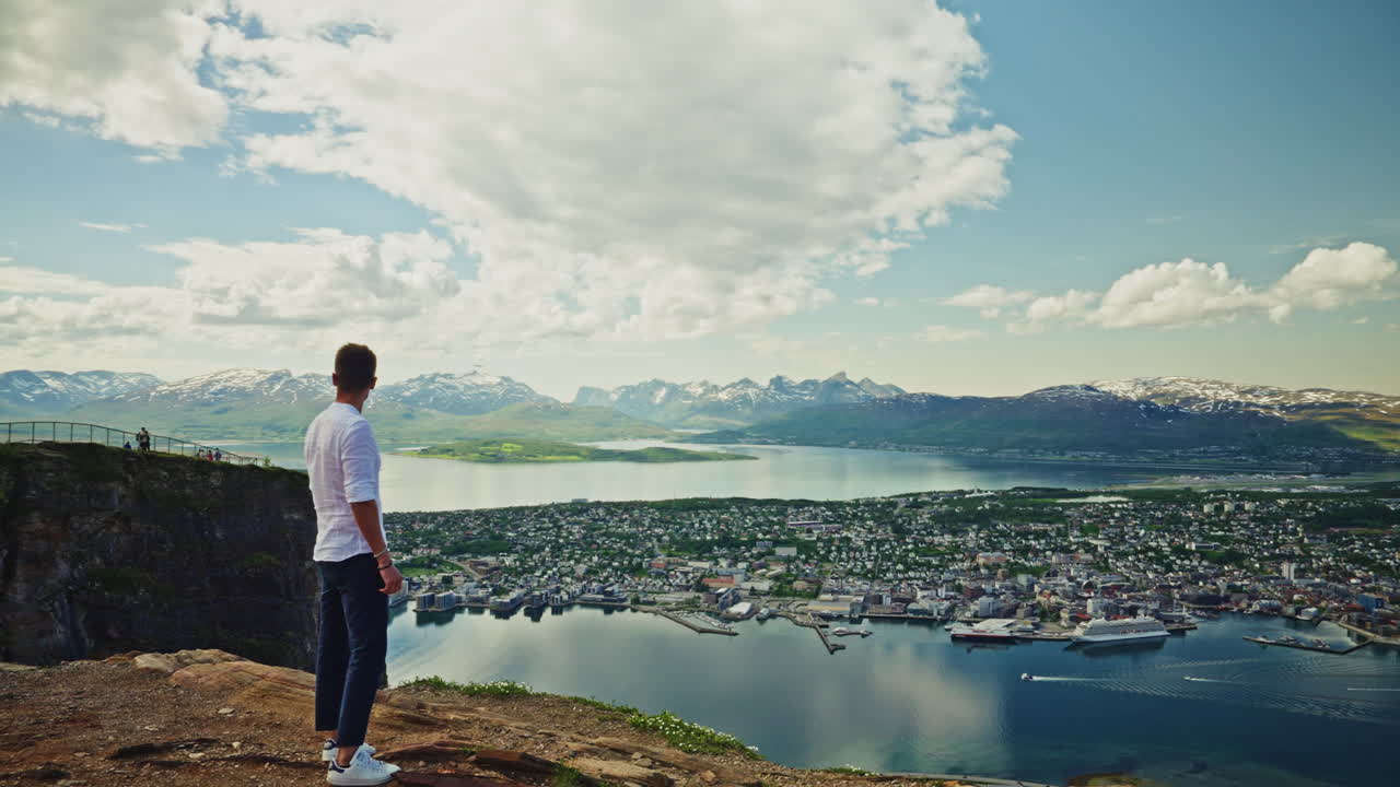 Young man with a white shirt enjoying the breathtaking view of Tromso from a high view point in Norway. High view of the nordic city, the landscape and the beautiful Norwegian fjords.