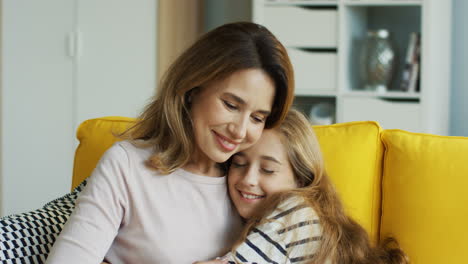 Close-Up-Of-Teenage-Blonde-Girl-Approaching-To-Her-Mother-Who-Is-Sitting-On-Yellow-Sofa-And-Hugging-Her