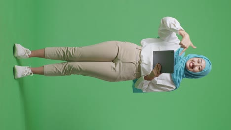 full body of asian muslim woman smiling and using tablet then pointing to it while standing in the green screen background studio