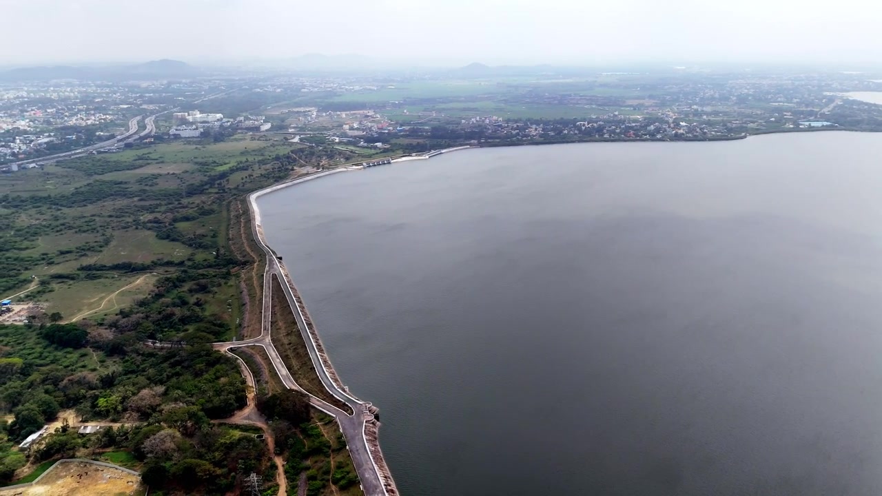 Ampio panorama del lago Chembarambakkam che mostra le calme acque del bacino, la caratteristica torre di presa, le infrastrutture costiere, i paesaggi aperti e lo skyline urbano in crescita di Chennai sotto un cielo sereno.