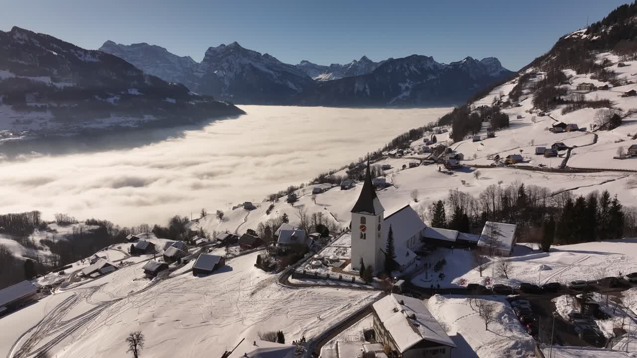 Aerial view of Amden in Glarus, Switzerland, with the church amidst snow-covered homes, while a sea of fog envelops Lake Walensee, framed by towering mountains.