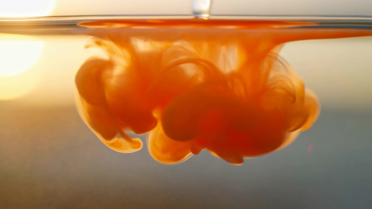 Nærbillede af undervandsbevægelse viser lyst orange pigment, der spreder sig under en glat horisontal vandlinje, danner en ekspanderende sky med pæreformede masser, fjerlignende filamenter, gennemsigtige striber og en varm glød