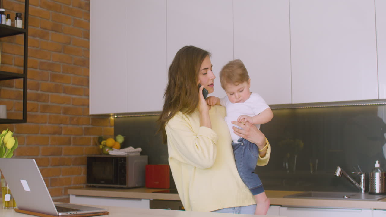 Woman Holding Her Baby In The Kitchen While Talking On The Phone 1