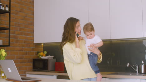 Woman-Holding-Her-Baby-In-The-Kitchen-While-Talking-On-The-Phone-1