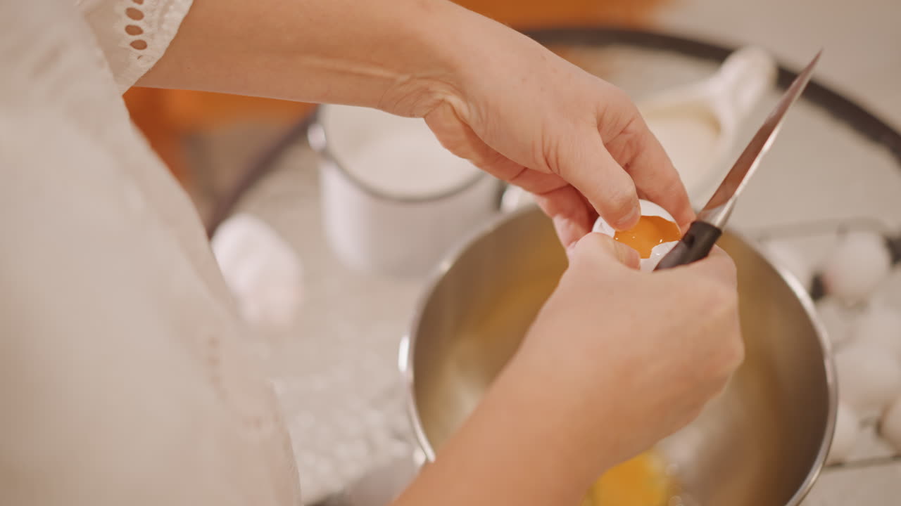 Overhead hands cracking egg into bowl, stainless mixing bowl and measuring cup visible, whisk at ready, multiple shells nearby, warm countertop, energetic prep for pumpkin pie batter and closeup food