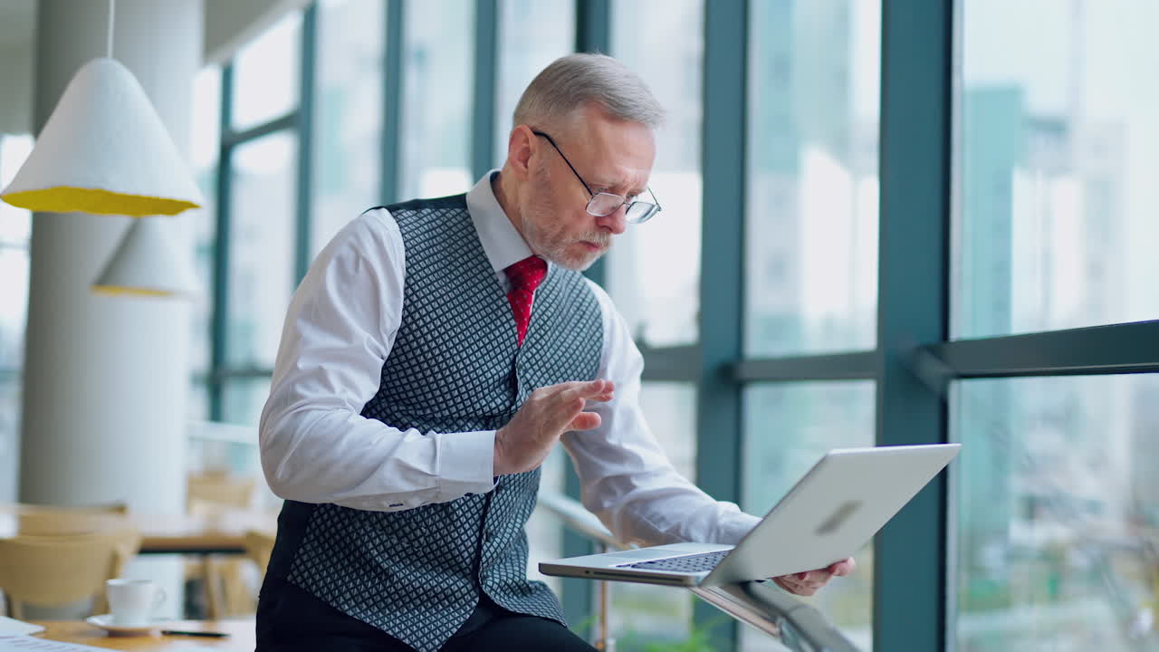 Nervous mature man working on a laptop. Serious senior businessman in eyeglasses standing near the window and looking at his laptop in his office.