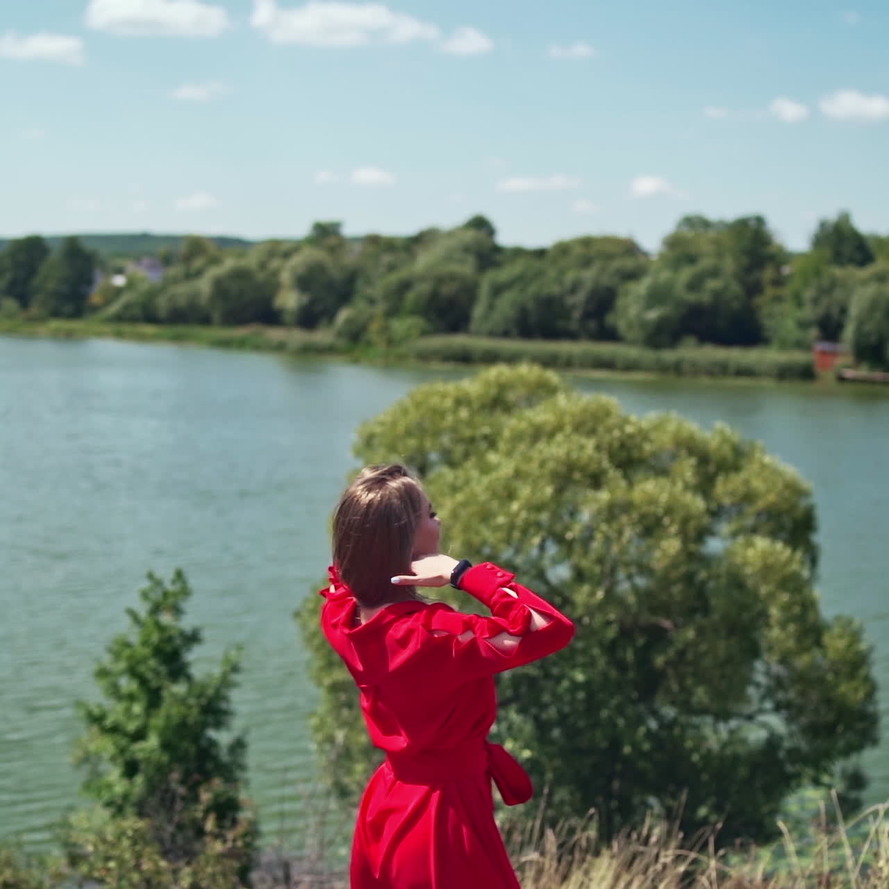 Backside view of pretty woman near the river. Young female in red dress feeling excited among nature in a summer day. Lady enjoys natural landscape. Slow motion.