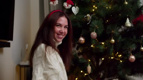 retrato de una feliz joven morena en un suéter blanco que sonríe y mira a la cámara cerca de su árbol de navidad decorado en una acogedora casa de invierno