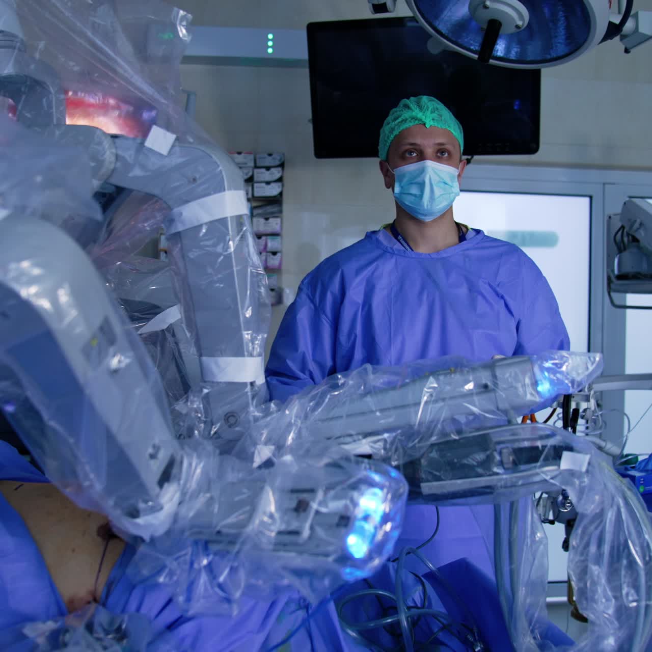 Futuristic robotic arms move very slowly in the belly of a patient. Male surgeon stands behind looking attentively on the monitors