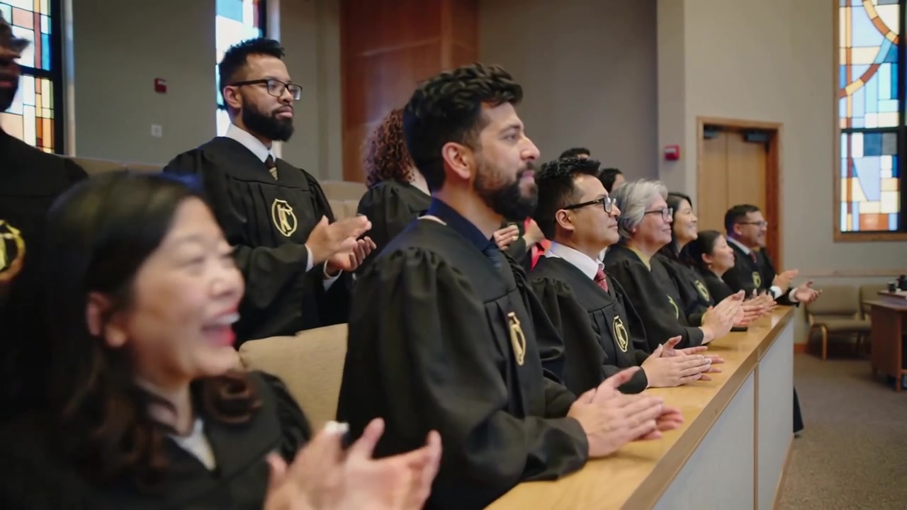 Applaus geactiveerd door een signaal van achter de schermen, een groep in gewaden staat op en applaudisseert bij een kerkbank, gouden schilden.