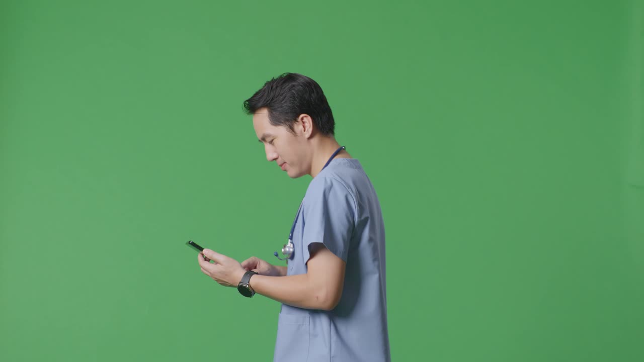 병원에서 녹색 화면 배경에서 는 동안 스마트폰을 사용하여 스테토스코프를 가진 아시아 남성 의사의 측면 시각