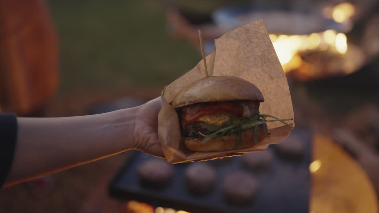 Hand holding a burger by a campfire