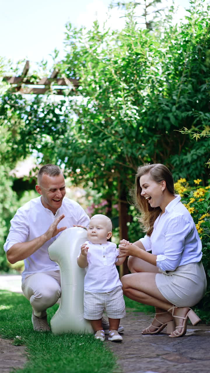 Cute little blond baby boy stands near his parents. Smiling dad shows number one balloon to his kid. First child's birthday. Vertical video.