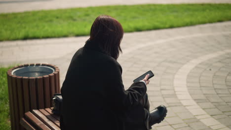 close-up of woman sitting on park bench, looking down at smartphone, deeply focused on screen with a relaxed posture, the background is a blurred view of greenery and a walking path