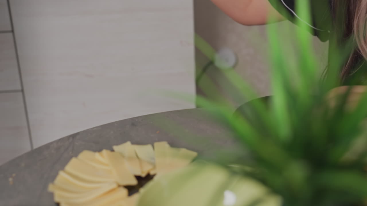 woman in green lounge outfit stands at kitchen counter placing sliced sausage and cheese on plate near wooden chopping board and kettle while preparing meal in cozy home