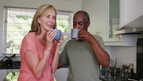 happy diverse senior couple drinking coffee and talking standing in kitchen