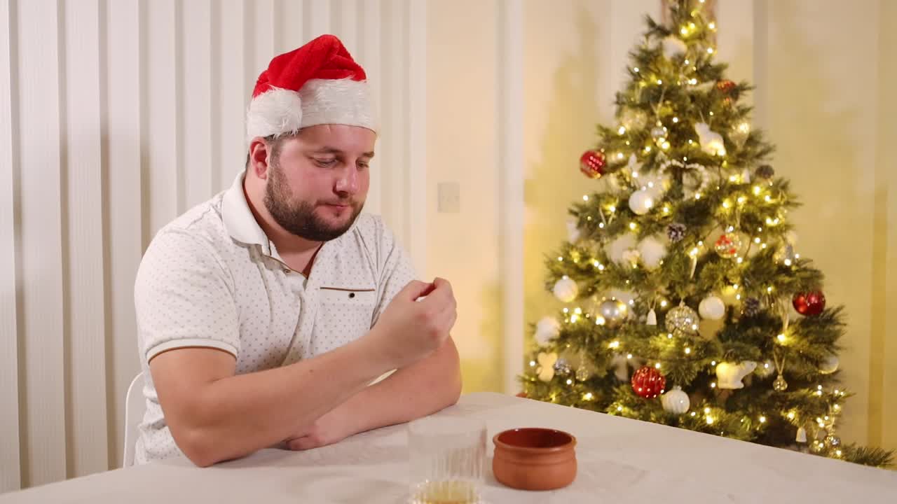 joven sentado al lado de un árbol de navidad decorado comiendo bocadillos de navidad mientras usa sombrero de santa, vista estática
