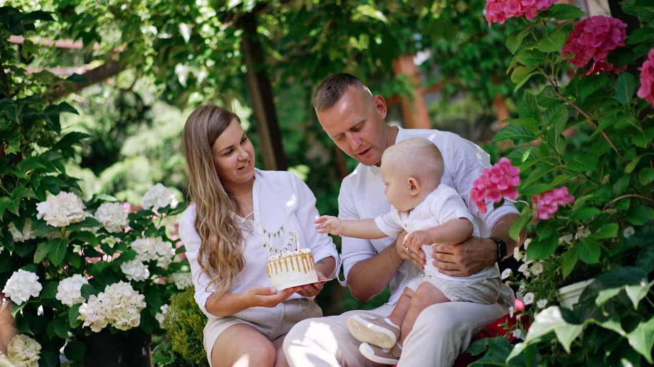 Beautiful young Caucasian family sitting in the blossoming garden. Baby boy sitting on dad's laps tries to get the birthday cake in mom's hands. First year of child's life.