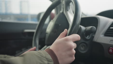 close up of fair skinned hand reaching toward steering wheel inside parked car with blurred modern city buildings in background on overcast winter day