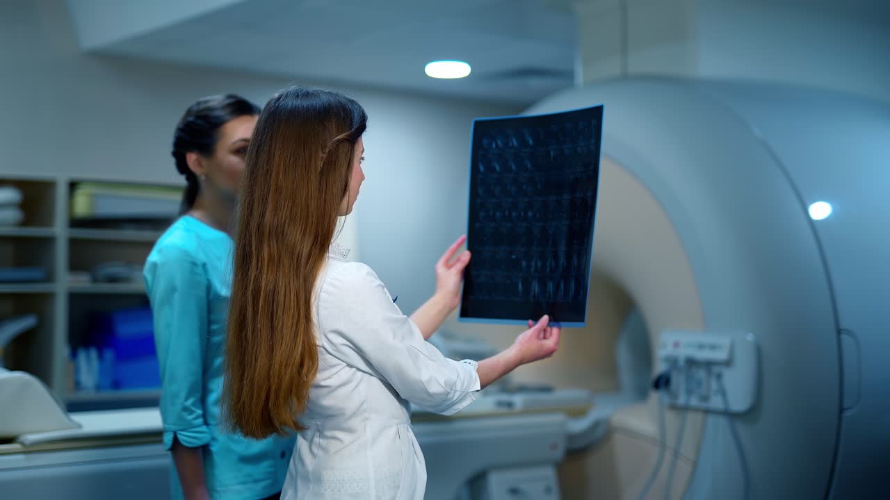 Female doctor and a nurse looking at x-ray photo. Two experts analyzing radiograph picture on the modern medical equipment background in clinic.