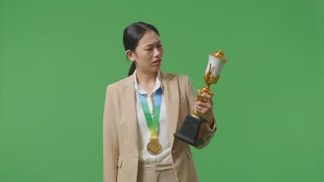 금메달을  슈트를 입은 아시아 사업가 여성이 트로피를 바라보고 머리를 흔들며 불만을 품으며 스튜디오의 녹색 스크린 배경에서 첫 번째 우승자로 승리합니다.