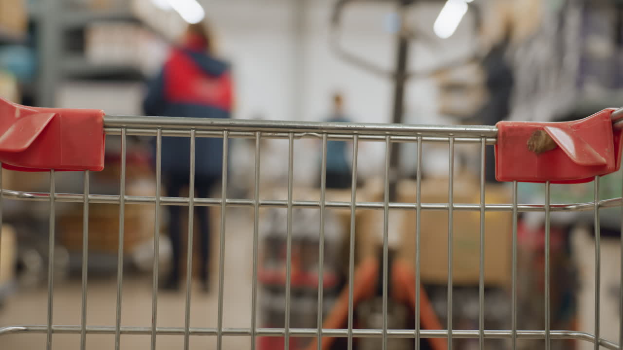 POV rolling wire cart glides down bright supermarket aisle while blurred shoppers push trolleys past stocked shelves, grid handle bars in sharp focus creating dynamic retail motion perspective