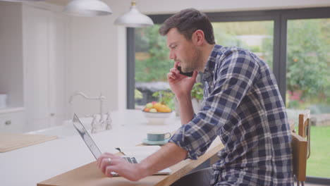 hombre trabajando desde casa usando una computadora portátil en el mostrador de la cocina hablando por teléfono móvil - filmado en cámara lenta