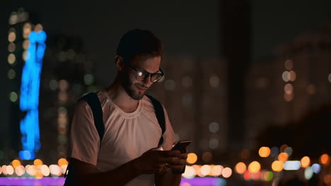 a young man with glasses at night looks at the smartphone screen and writes text messages for his blog on social networks. reads information and makes a trip martour