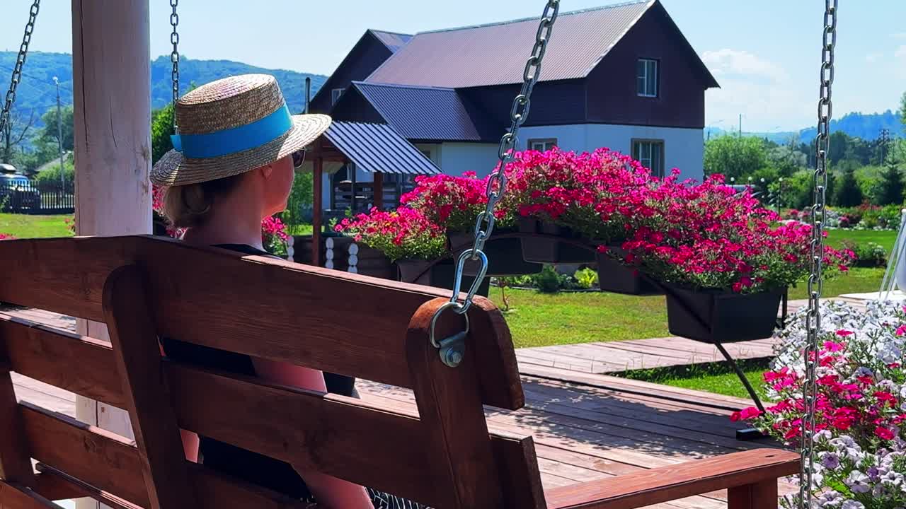 Serene Moments: A Woman in a Straw Hat Enjoys a Peaceful View of Colorful Flowers and a Charming House from Her Swing, Embracing Tranquility in Nature