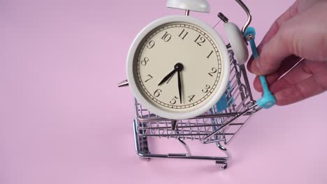 hand rolls a decorative supermarket trolley with a white alarm clock inside on a pink background