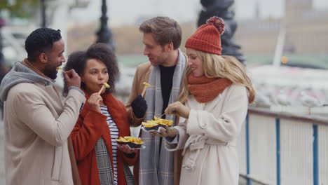 grupo de amigos al aire libre con abrigos y bufandas comiendo papas fritas para llevar en un viaje de otoño o invierno a londres - filmado en cámara lenta