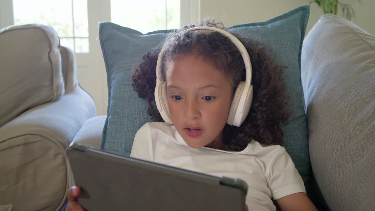 Female child reacting to surprising moment on tablet while sitting with blue cushion and headphones