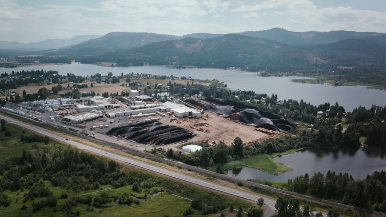Aerial timelapse video of Priest River, Idaho, shows an industrial lumber yard with stacked timber piles beside the river, mountains in the background, and a highway cutting through the green valley