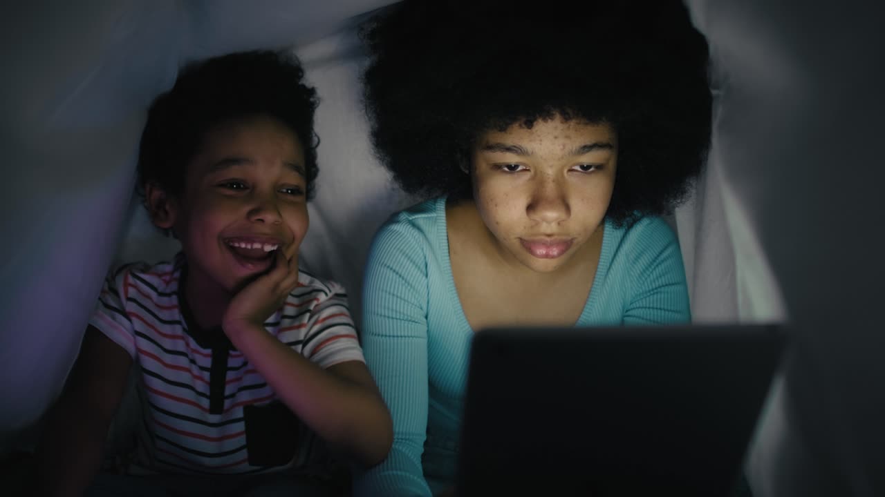 Two children browsing digital tablet under blanket at night