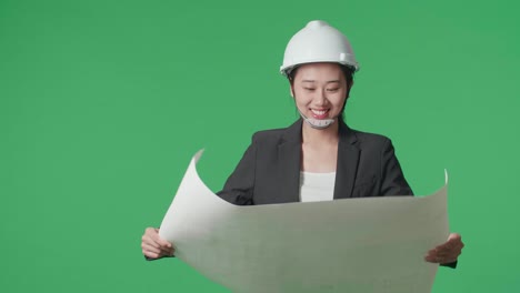smiling asian female engineer with safety helmet looking at blueprint while walking in the green screen studio