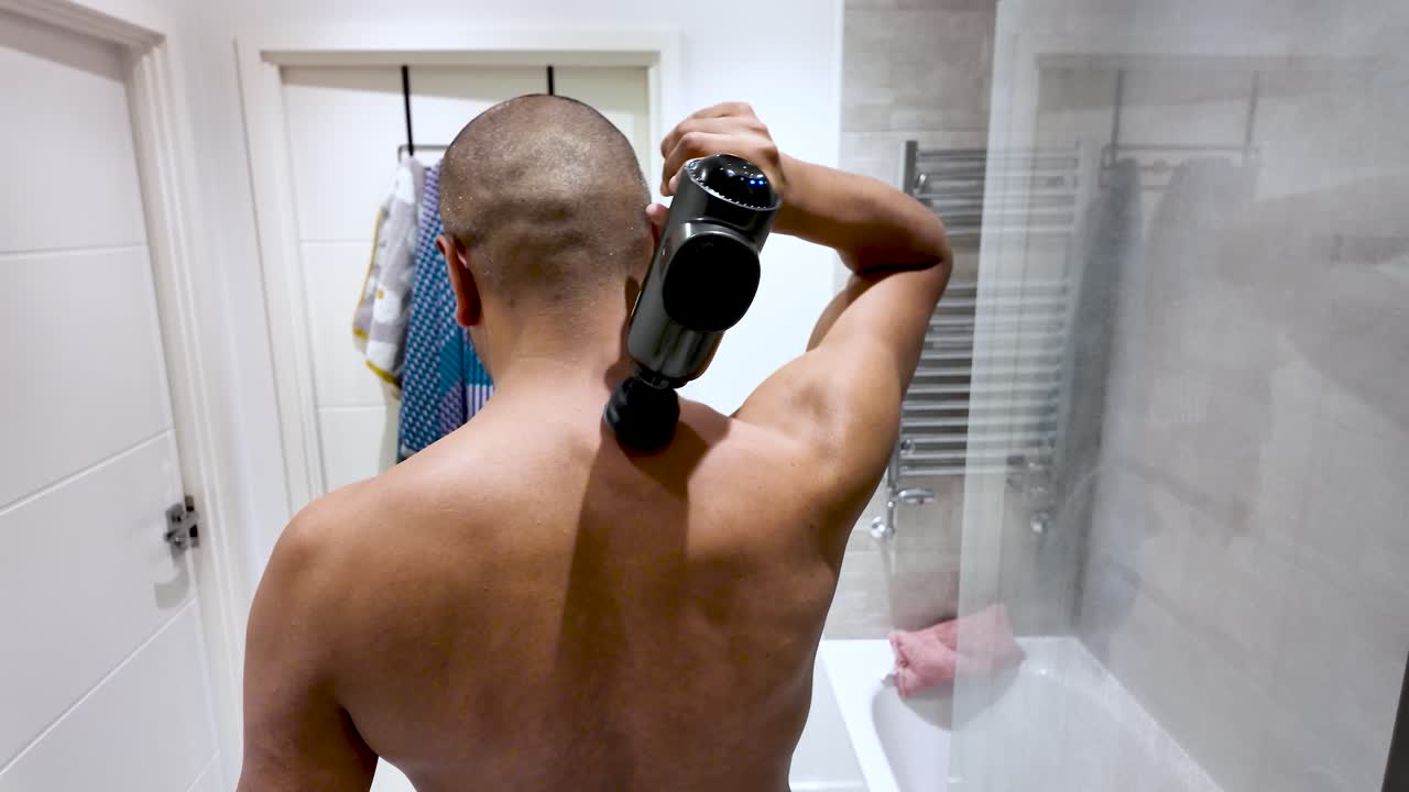 Shirtless man massaging his back with percussion massage gun in bathroom