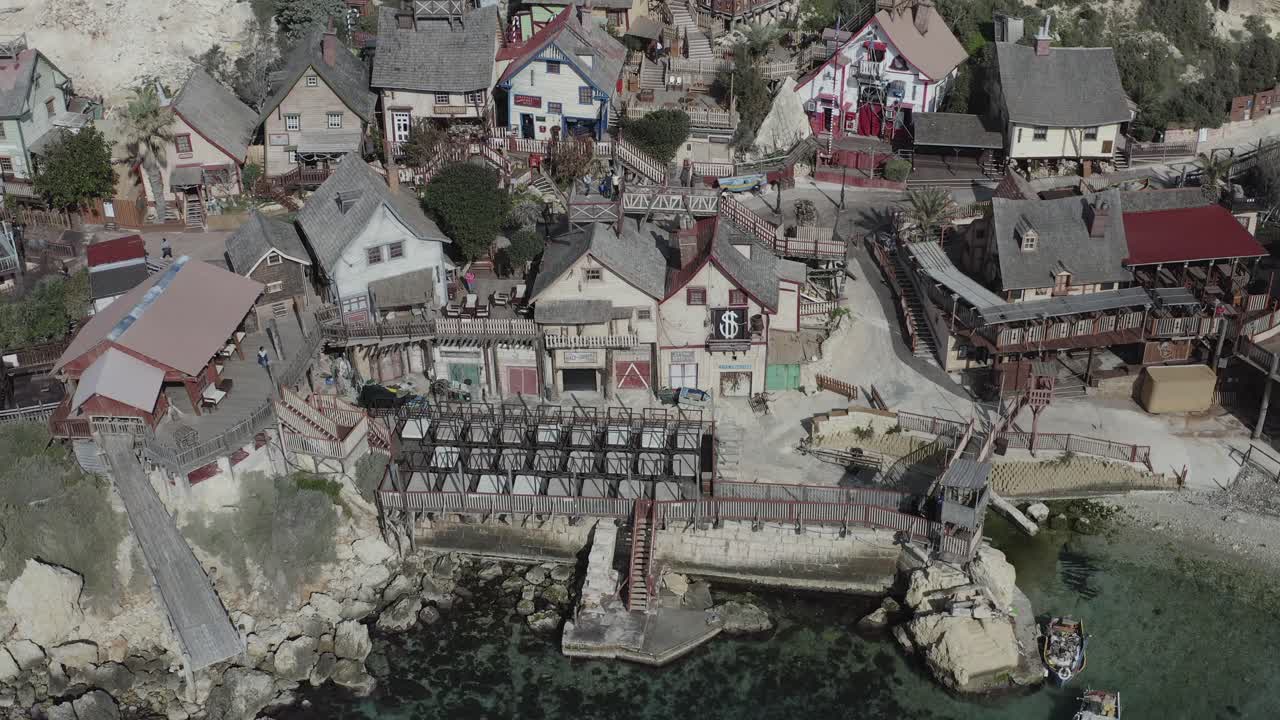 A drone gently pulls back from Popeye Village, revealing the cluster of colorful houses, Anchor Bay’s turquoise waters, and Malta’s picturesque coastline from a widening perspective