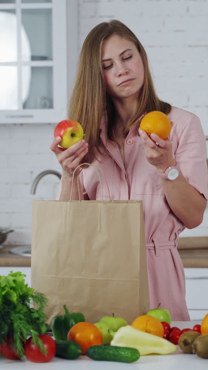 Pretty woman takes fresh fruit from paper bag. Young model is sniffing organic fruit on the light kitchen background. Many healthy food on a table for losing weight. Vertical video