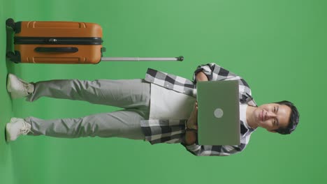 cuerpo lleno de viajero masculino asiático con equipaje usando una computadora portátil luego sonriendo a la cámara mientras está de pie en el estudio de fondo de pantalla verde