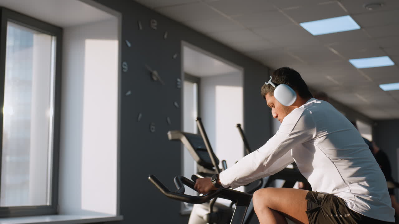 Student in white activewear cycles on stationary bike wearing headphones, while others run on treadmills and lift weights around him in bright gym environment with large windows and modern equipment