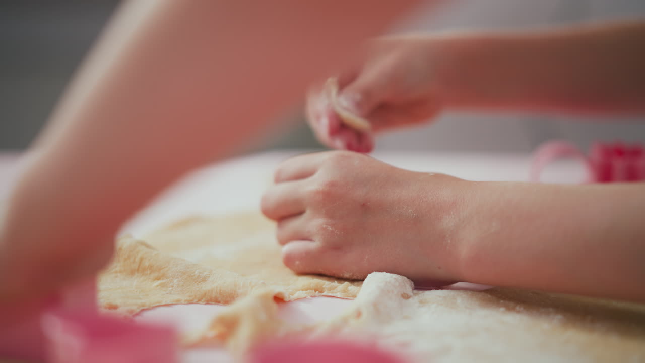 Close up of child twisting dough with pink shape cutter while another child rubs flour on dough in hand, soft warm lighting capturing playful baking moment during kitchen activity