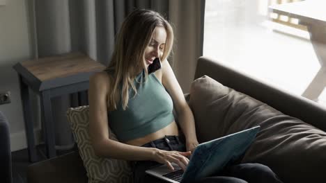 Blonde-woman-sitting-on-sofa-with-laptop-and-talking-by-smartphone